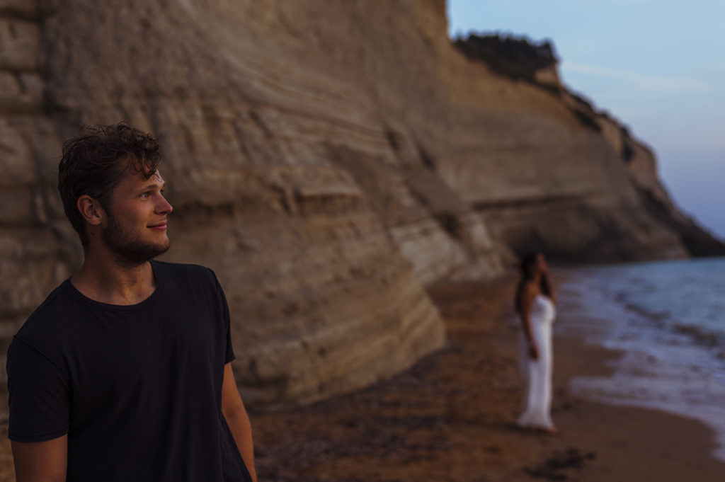 Greece Destination Engagement - Corfu - Boyfriend staring at the sunset, girlfriend in the background